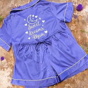 May include: A lilac satin pajama set with white trim, featuring a collared, short-sleeved top and matching shorts. The top is embroidered with a moon, hearts, stars, and the words "Sweet dreams, Mom."