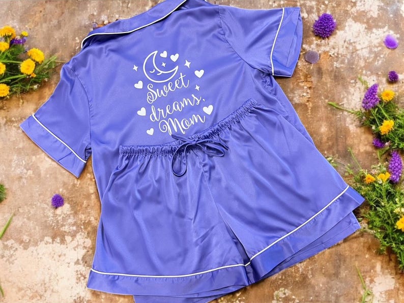 May include: A lilac satin pajama set with white trim, featuring a collared, short-sleeved top and matching shorts. The top is embroidered with a moon, hearts, stars, and the words "Sweet dreams, Mom."