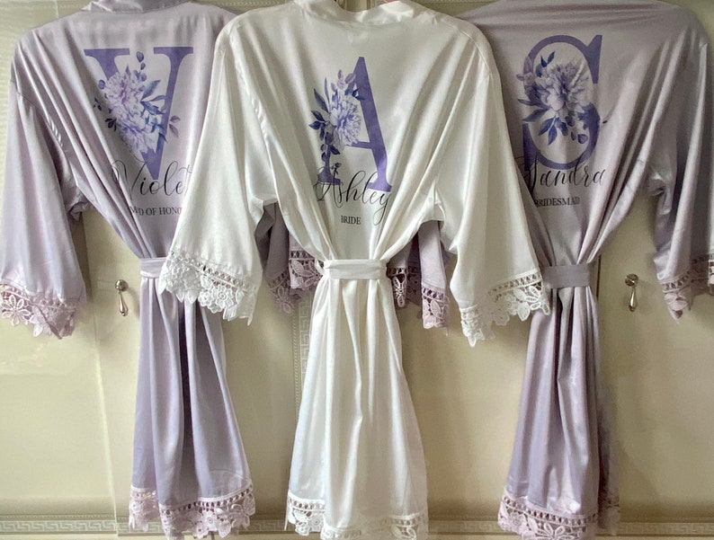Peut inclure: Trois robes en satin avec bordure en dentelle, une blanche et deux lavande, avec des monogrammes floraux personnalis&eacute;s et du texte. La robe blanche dit "Ashley Bride". Les robes lavande disent "Violet Maid of Honor" et "Sandra Bridesmaid".
