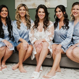 Puede incluir: Cinco mujeres en una foto de grupo. Cuatro mujeres llevan batas de sat&eacute;n azul claro y una mujer lleva una bata de encaje blanco. Las mujeres est&aacute;n sentadas en un sof&aacute; gris.