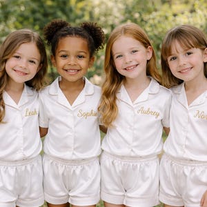 May include: Four young girls wearing matching ivory pajama sets with gold embroidered names. The sets include short-sleeved button-down shirts and shorts. The girls are smiling, posing in front of a blurred green background.