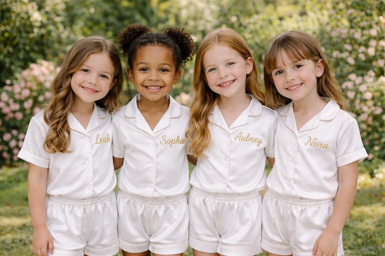 May include: Four young girls wearing matching ivory pajama sets with gold embroidered names. The sets include short-sleeved button-down shirts and shorts. The girls are smiling, posing in front of a blurred green background.
