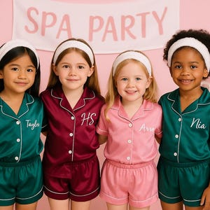 May include: Four young girls wearing matching satin pajama sets in teal, burgundy, and pink, with white piping. Each set is personalized with a name embroidered on the shirt. They are wearing white headbands, posing in front of a pink backdrop with a "SPA PARTY" banner.
