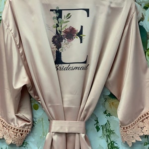Peut inclure: Une robe de chambre en satin rose poudr&eacute; avec un motif floral et la lettre "E" en noir. La robe a des bordures en dentelle sur les manches et dit "bridesmaid" au dos.