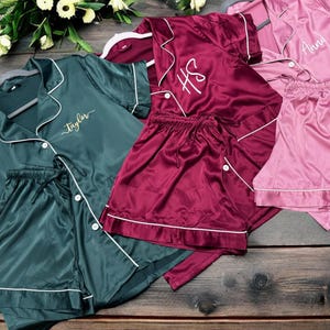 May include: An assortment of satin pajama sets in shades of teal, burgundy, and pink, with white piping and personalized embroidery. The sets include button-down shirts and matching shorts, displayed on a wooden surface.