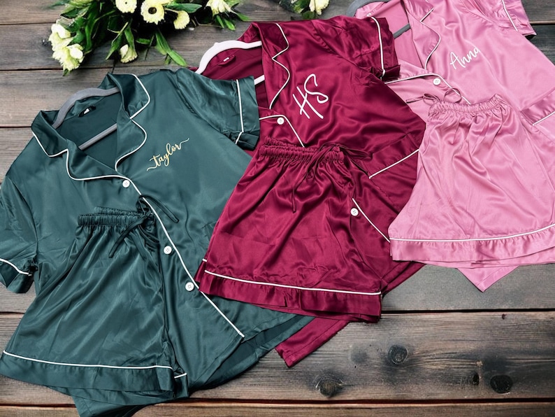 May include: An assortment of satin pajama sets in shades of teal, burgundy, and pink, with white piping and personalized embroidery. The sets include button-down shirts and matching shorts, displayed on a wooden surface.
