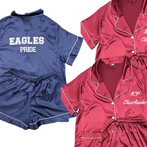 May include: Navy blue satin pajama set with white trim. The top has "EAGLES PRIDE" embroidered on the back. Two maroon satin pajama sets with white trim are also shown, one with "Xaverian Cheerleader" and the other with "KP Cheerleader" embroidered.