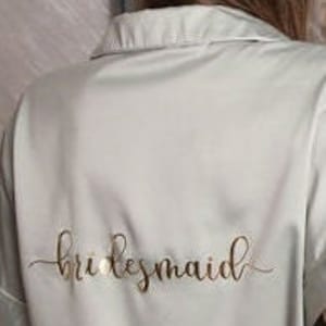 Puede incluir: Una bata de sat&eacute;n verde claro con la palabra "bridesmaid" escrita en letras doradas en la espalda.
