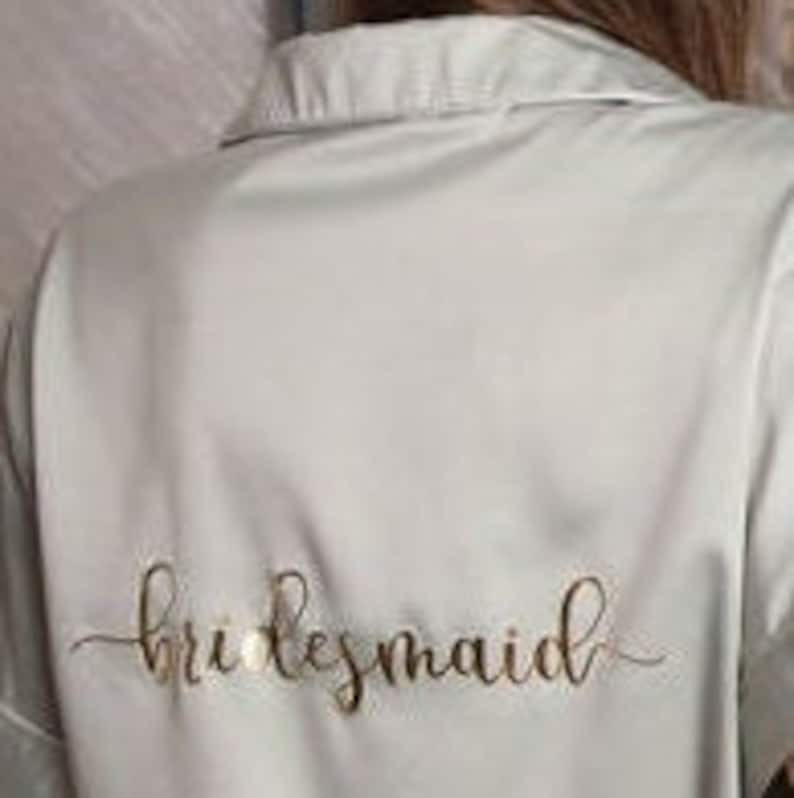 Puede incluir: Una bata de sat&eacute;n verde claro con la palabra "bridesmaid" escrita en letras doradas en la espalda.