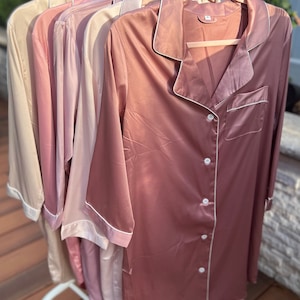 Puede incluir: Cinco camisas de dormir de sat&eacute;n en varios tonos de rosa, colgadas de perchas. Las camisas tienen un frente abotonado y un cuello con ribete blanco.