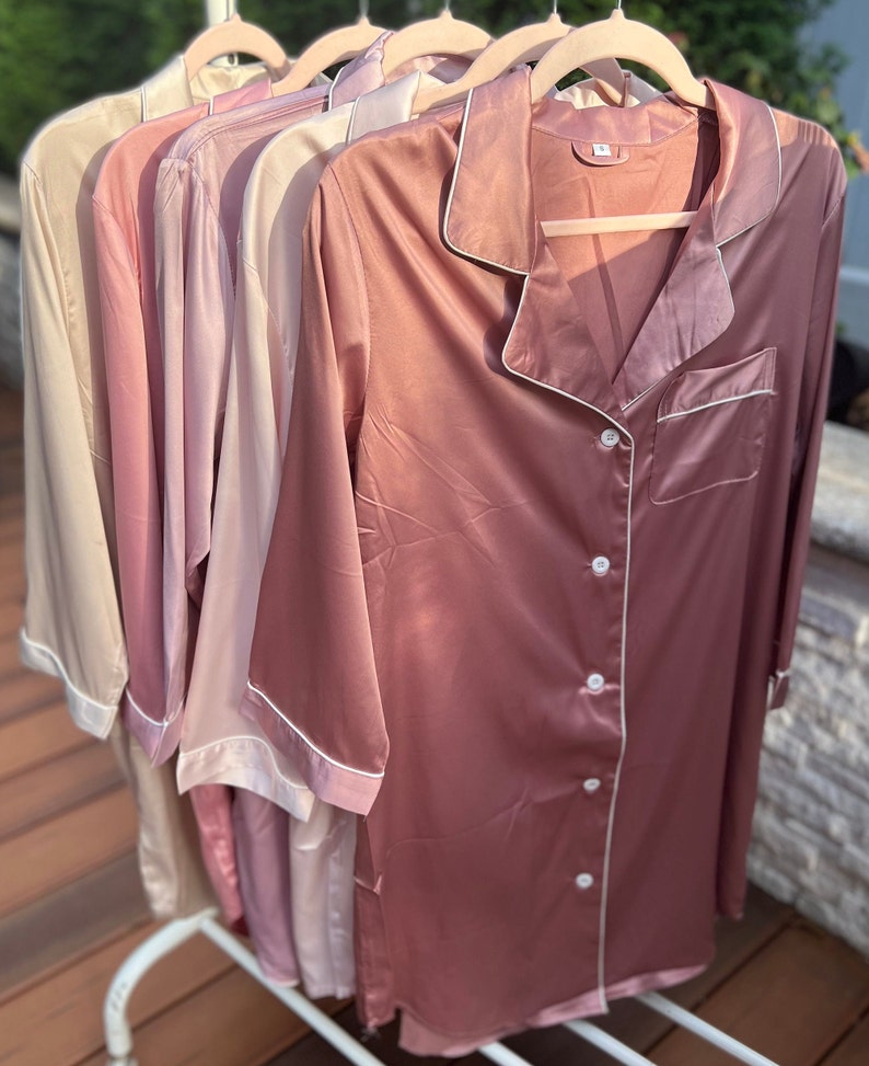 Puede incluir: Cinco camisas de dormir de sat&eacute;n en varios tonos de rosa, colgadas de perchas. Las camisas tienen un frente abotonado y un cuello con ribete blanco.