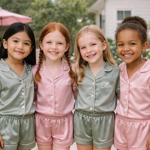 May include: Four young girls wearing matching satin pajama sets in sage green and pink. Each set includes a short-sleeved button-down shirt and shorts, with names embroidered on the shirts. The girls are smiling in front of a pool.