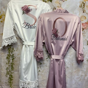 Peut inclure: Deux robes en satin avec des bordures en dentelle. La robe blanche a un "S" dor&eacute; avec des accents floraux et le texte "Bride". La robe rose a un "O" dor&eacute; avec des accents floraux et le texte "Bridesmaid".