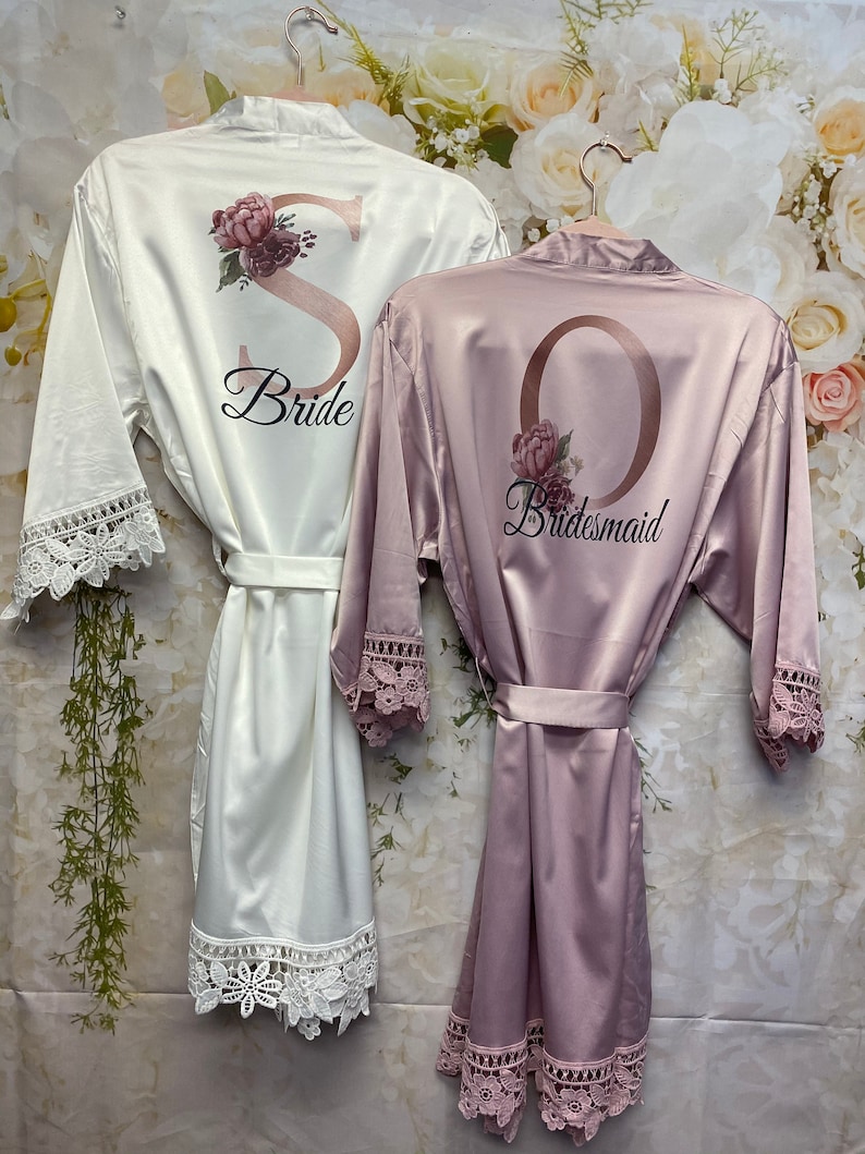 Peut inclure: Deux robes en satin avec des bordures en dentelle. La robe blanche a un "S" dor&eacute; avec des accents floraux et le texte "Bride". La robe rose a un "O" dor&eacute; avec des accents floraux et le texte "Bridesmaid".