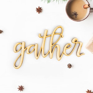 May include: Wooden sign with the word "gather" in a cursive font. The sign is on a white background with pine branches, cinnamon sticks, and star anise.
