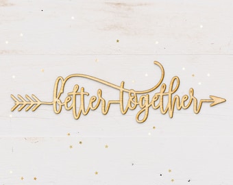 May include: Wooden sign with the words "better together" in a cursive font, with an arrow on each side. The sign is on a white wood background with gold glitter.