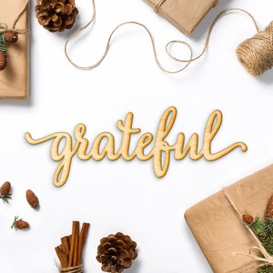 May include: Wooden sign with the word "grateful" written in cursive script. The sign is surrounded by brown paper packages, pine cones, cinnamon sticks, and twine.