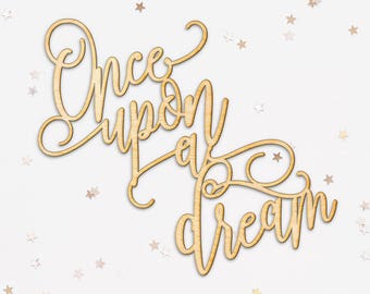 May include: Wooden cutout in a cursive font that reads "Once upon a dream." The cutout is light brown and surrounded by small star-shaped confetti on a white background. Ideal for crafts and decor.