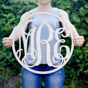 May include: A round wooden sign with the letters "ARE" cut out in a decorative script font. The sign is held by a person in front of a green leafy background.
