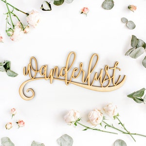 May include: A wooden sign with the word "wanderlust" written in cursive script. The sign is surrounded by pink flowers and green leaves on a white background.