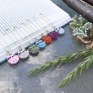 May include: A set of eight gold bookmark clips with colourful felt circles attached. The circles have a leopard print design with black spots and pink glitter accents. The colours of the circles are pink, grey, green, blue, orange, purple, and red.