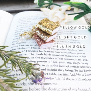 Peut inclure: Un collier doré avec trois breloques miniatures en forme de livre, en or jaune, or clair et or rosé pailleté. Les breloques sont empilées. Le collier est présenté sur un livre ouvert avec du texte.