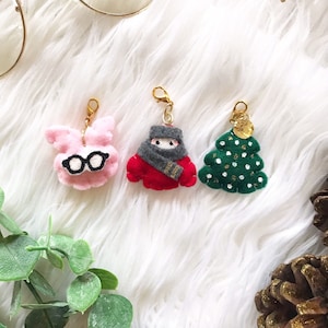 May include: Three handmade felt charms: a pink bunny wearing glasses, a red-clad figure with a gray scarf, and a green Christmas tree with gold accents. Each charm has a gold-colored clasp.