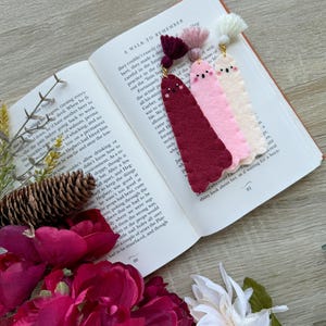 Könnte beinhalten: Drei Filz-Lesezeichen-Ornamente mit gestickten Gesichtern und Wollquasten. Die Lesezeichen sind burgunderrot, rosa und cremefarben.