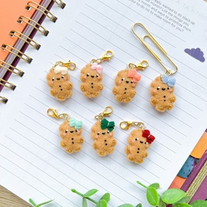 May include: Seven felt gingerbread person charms with different colored bows. The charms are attached to gold lobster clasps. The charms are on a white lined notebook.