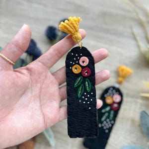 May include: Two black felt bookmarks with embroidered flowers in shades of pink, red, yellow, and green. Each bookmark has a gold tassel at the top.