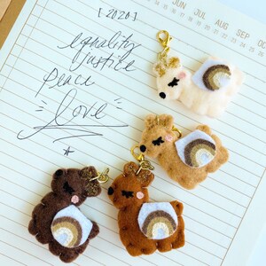 May include: Four felt llama keychains with rainbow accents. The llamas are brown, tan, and white. The keychains are attached to gold rings.