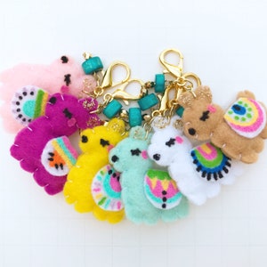 May include: A collection of colorful felt llama keychains. Each keychain features a different color llama, including pink, purple, yellow, and blue. The llamas have decorative saddle blankets and gold-colored clasps with turquoise beads.