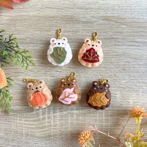 Op de afbeelding: Zes vilten sleutelhangers in de vorm van beren met goudkleurige sluitingen. De beren zijn in verschillende kleuren, waaronder wit, bruin en beige, en hebben blad- en pompoenontwerpen. De sleutelhangers zijn gerangschikt op een houten oppervlak met herfstbladeren.