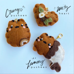 Peut inclure: Trois porte-clés Pusheen en feutre marron avec des fermoirs dorés. Un porte-clés a un arc-en-ciel, un autre a des fleurs et le troisième a un motif blanc et marron. Les mots "Courage", "Unity" et "Harmony" sont écrits au-dessus et en dessous.