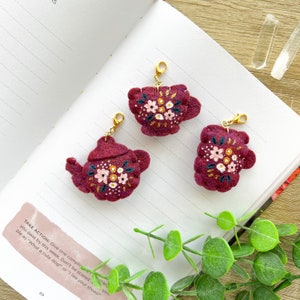 Peut inclure: Trois breloques en feutre en forme de tasses à thé et de théières, avec des détails floraux brodés en fil rose, blanc et or. Les breloques sont de couleur rouge bordeaux et ont des fermoirs en métal doré.