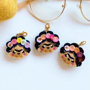 May include: Three felt keychains featuring a stylized portrait of Frida Kahlo with a flower crown. The keychains are brown, black, and pink.