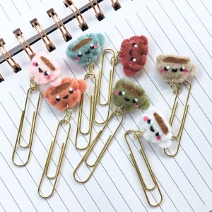 May include: A collection of handmade paper clips with felt coffee cup designs in various colors, including pink, brown, green, and white. The paper clips are gold-colored and attached to a spiral notebook.