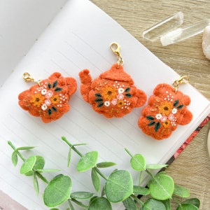 Op de afbeelding: Drie vilten theepotten met geborduurde bloemenmotieven in tinten van oranje, roze en groen. Elke theepot heeft een gouden metalen lus.