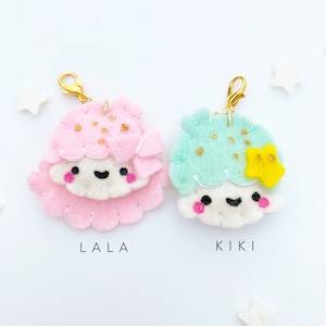 May include: Two felt character charms with gold clips. The charm on the left is pink with gold glitter and says "LALA". The charm on the right is light blue with gold glitter and says "KIKI".