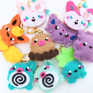 May include: A collection of felt keychains featuring various cartoon characters. The keychains are colorful and include characters like a pink and white bunny, a purple and blue creature, a brown and pink character, and a green and blue character.