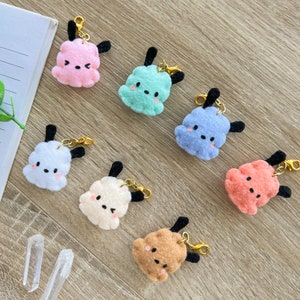 Peut inclure: Six breloques en peluche de différentes couleurs, dont rose, bleu, vert, blanc, beige et marron. Chaque breloque présente un visage de dessin animé avec des yeux noirs et une petite bouche. Les breloques sont attachées à des porte-clés dorés.