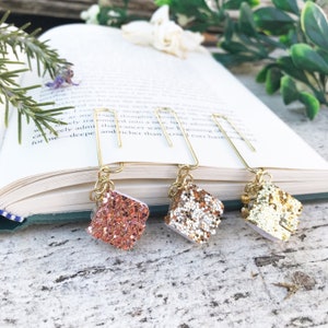 May include: Three gold book clips with glitter covered charms. The charms are shaped like small books and are in shades of pink, gold, and white.