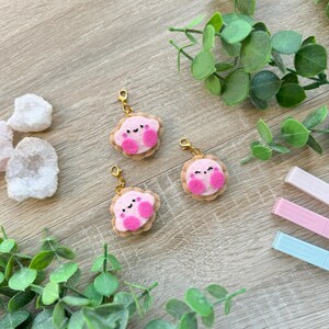 Op de afbeelding: Drie roze vilten bedels in de vorm van een personage met een glimlachend gezicht en twee kleine voetjes. De bedels zijn bevestigd aan gouden sleutelhangers.