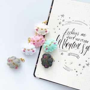 May include: Five felt cloud keychains in different colors, including white, pink, light blue, gray, and black. Each cloud has a happy face embroidered on it with gold thread. The keychains are arranged on a white surface next to a notebook with the text "feelings are just visitors let them come & go" written in black ink.