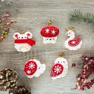 May include: Five felt Christmas ornaments with gold accents. The ornaments are a white bear wearing a red scarf, a red and white mushroom, a white swan with a red and white snowflake, a white owl with a red and white snowflake, and a white elephant with a red and white snowflake.