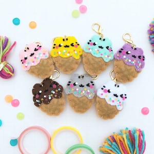 May include: Seven felt cupcake keychains with different colored frosting and sprinkles. The cupcakes are brown with white stitching around the edges. Each cupcake has a gold keychain attached.