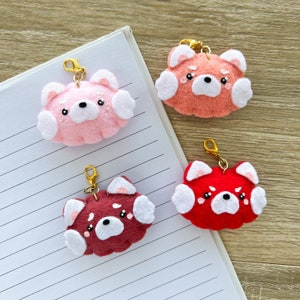 Peut inclure: Quatre breloques en feutre en rose, pêche, bordeaux et rouge. Chaque breloque présente un motif en forme d'ours avec des accents blancs et un fermoir doré. Les breloques sont disposées sur une page de carnet lignée et une surface en bois.
