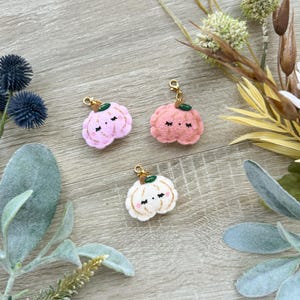 Op de afbeelding: Drie vilten pompoencharms met gouden accenten. De pompoenen zijn roze, perzik en wit met een schattig gezicht en glitterdetails.