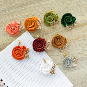 Op de afbeelding: Tien viltbloemencharms met gouden glitteraccenten. De bloemen zijn in verschillende kleuren, waaronder rood, oranje, roze, wit en groen. Elke bloem heeft een gouden lus om te bevestigen aan een sleutelhanger, ketting of ander knutselproject.