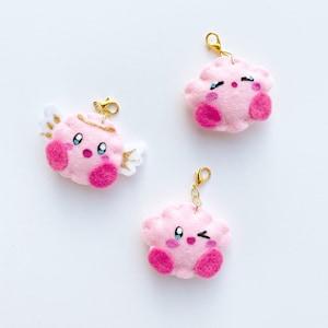 May include: Three pink felt keychain charms shaped like a cartoon character with different facial expressions. One charm has a halo and angel wings, one is winking, and one has its eyes closed.